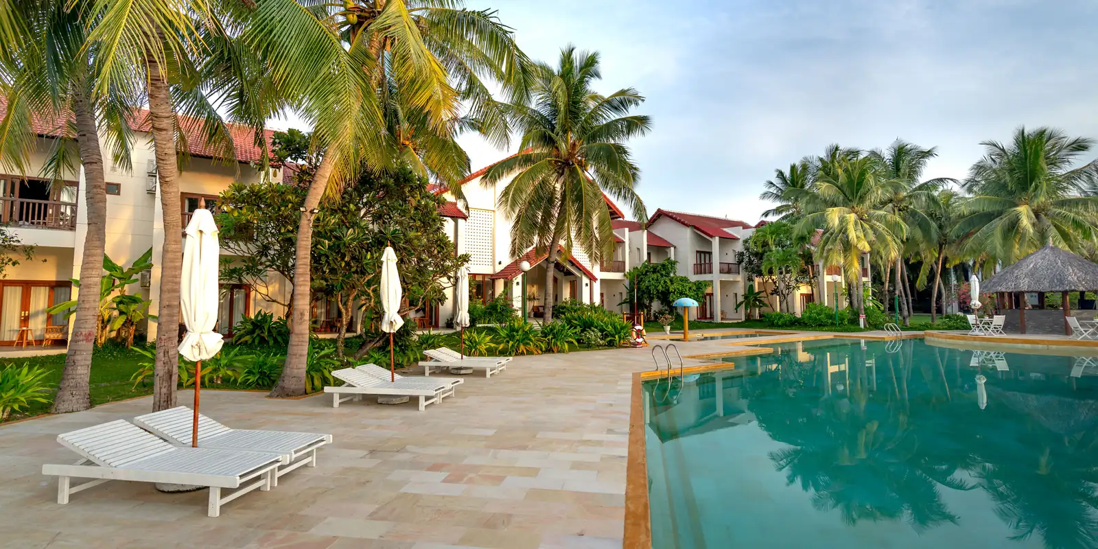 Stone-paved terrace with sun loungers, coconut palms and a pool beside low villas